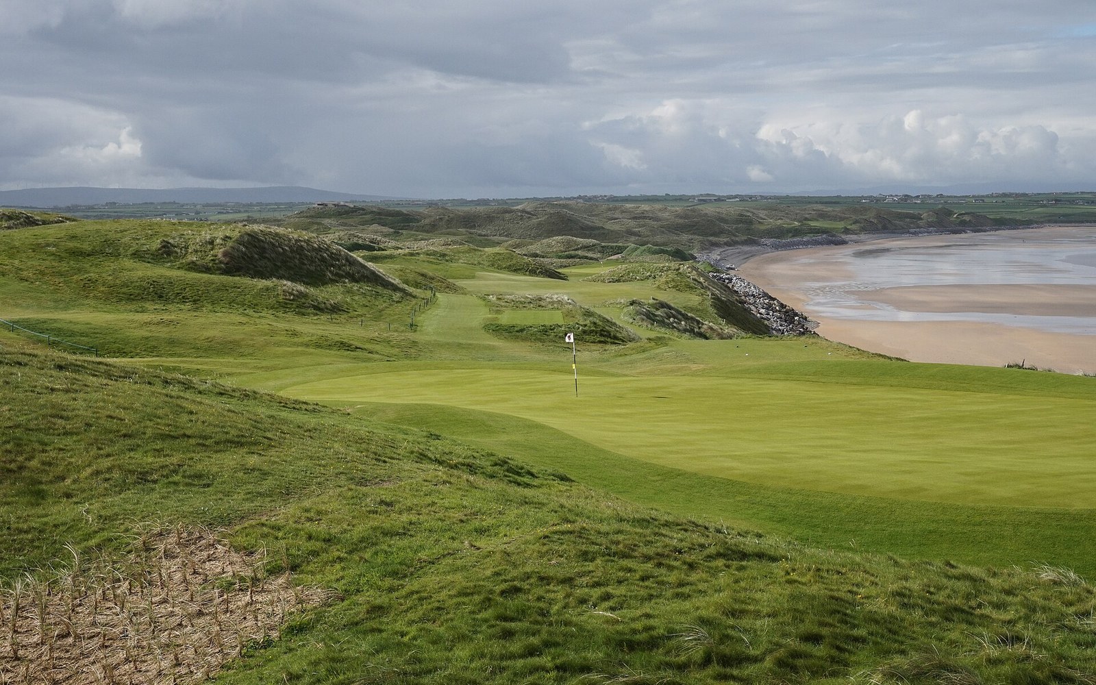 Dune toe at Ballybunion Old Course, sand face cut back below the marram overhang, adult greenkeeper standing at the break for scale, flat Atlantic light