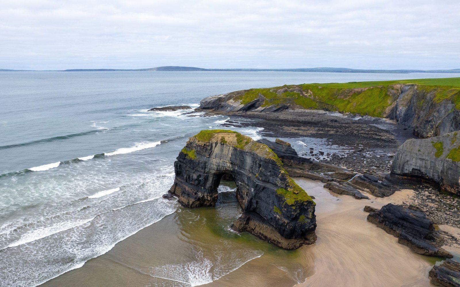 A grass-topped sea arch rises from a sandy beach along a rocky coastline