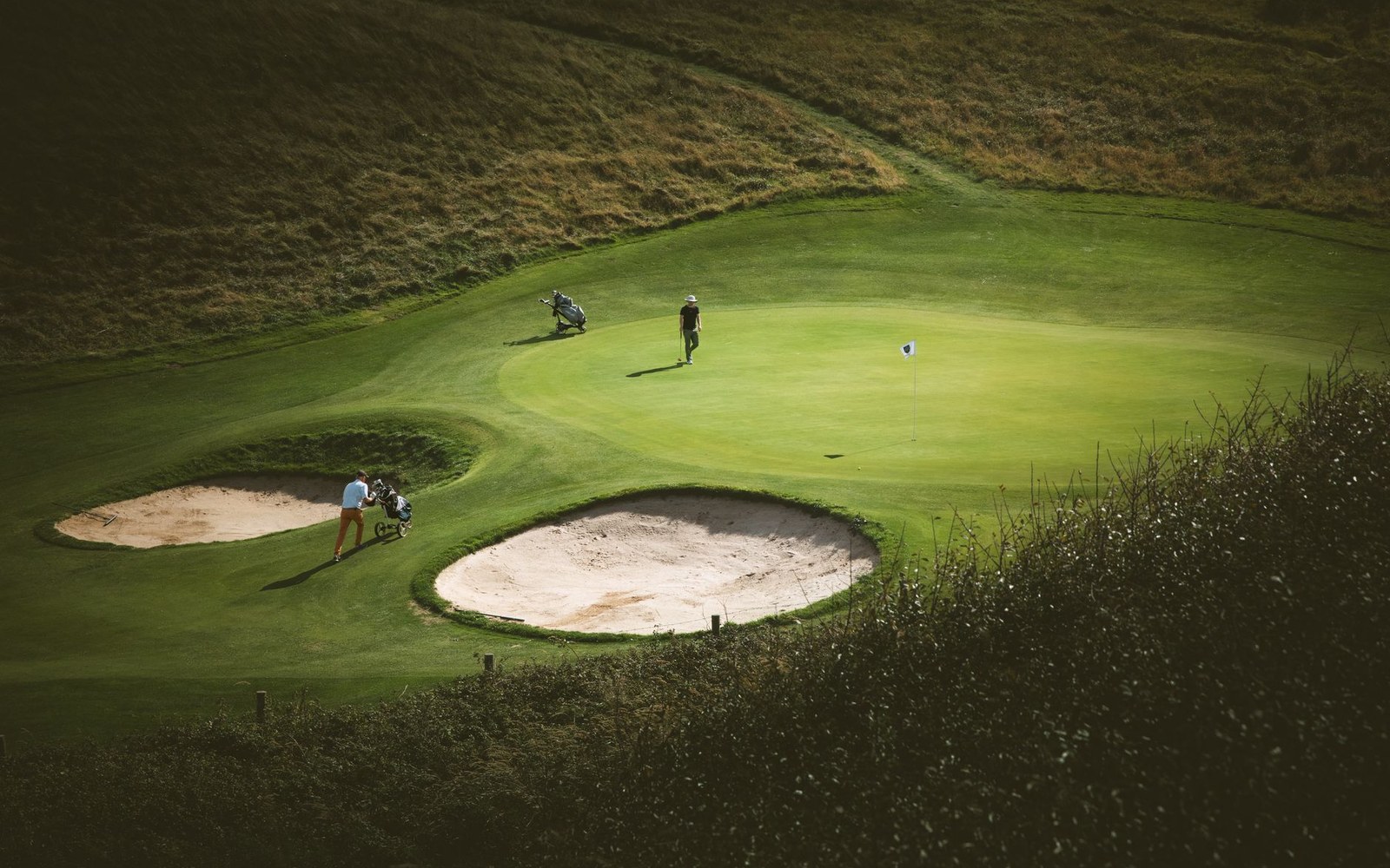 Golfers approach a green flanked by sand bunkers on a hilly links course