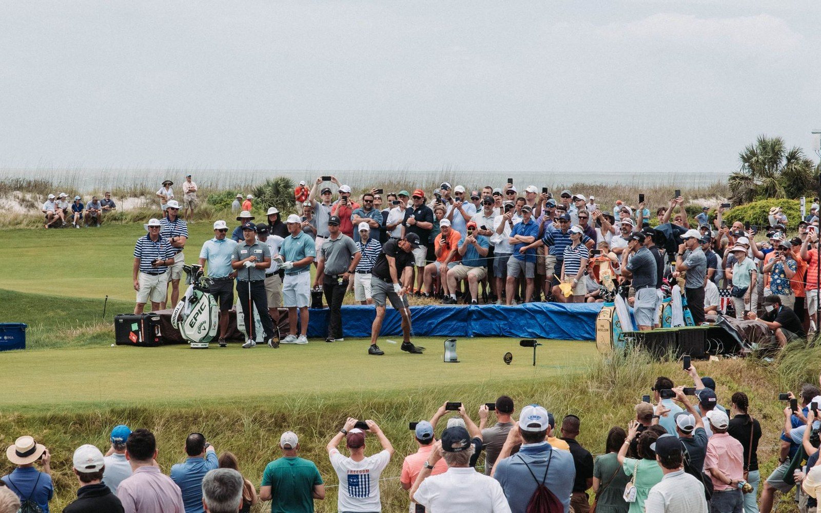 A golfer swings on a coastal green surrounded by a dense crowd of spectators