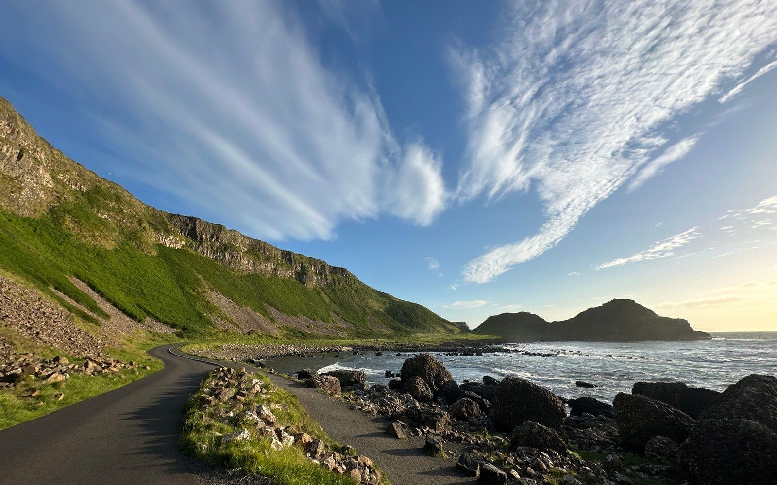 Coastal path winding along green cliffs beside a rocky shoreline under wispy clouds