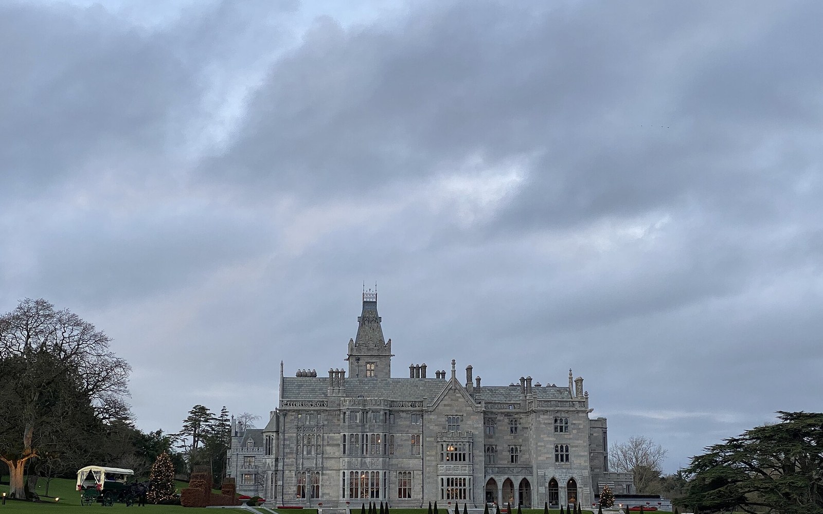 Gothic-style manor house with turrets stands beneath a cloudy sky, surrounded by trees