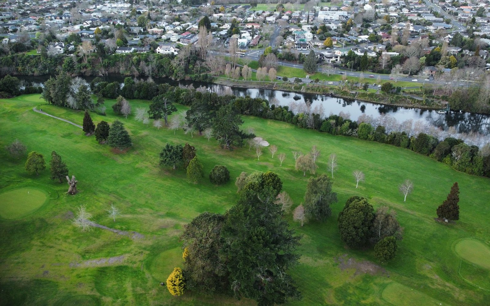 Aerial view of a golf course lined with trees beside a river and residential suburb